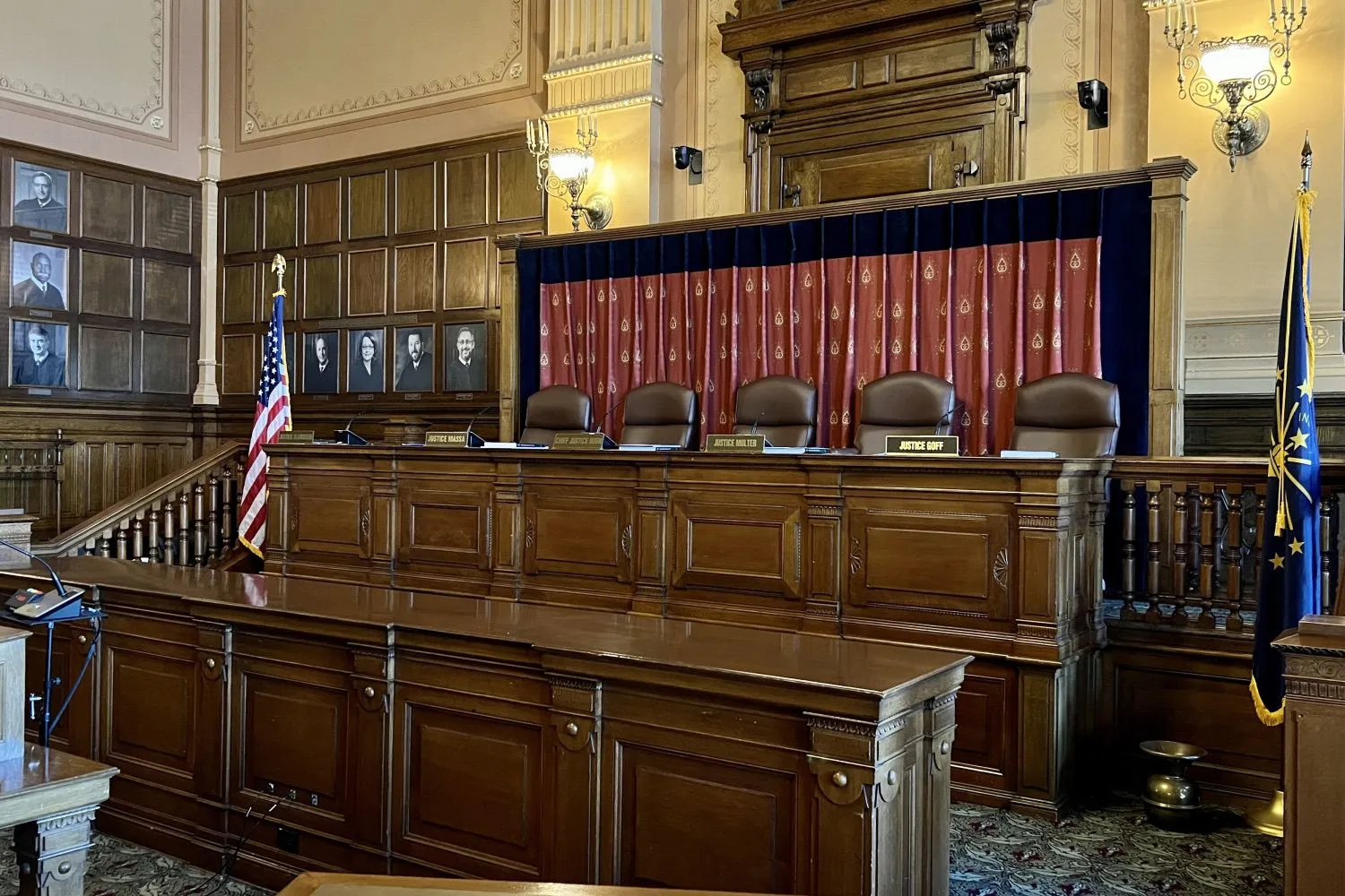 Wood-paneled Indiana courtroom with the justices' bench, the state and U.S. flags, and portraits of former justices along the wall.